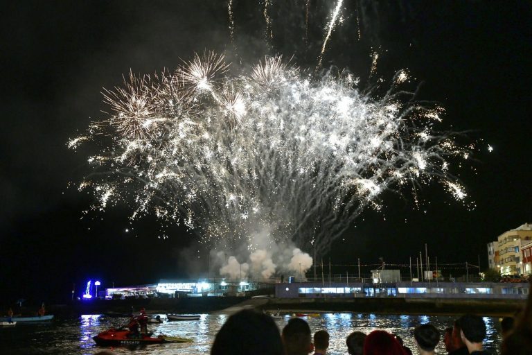 Fuegos artificiales blancos estallan de noche sobre el agua mientras el público mira de espaldas y una moto de agua de rescate espera en la orilla