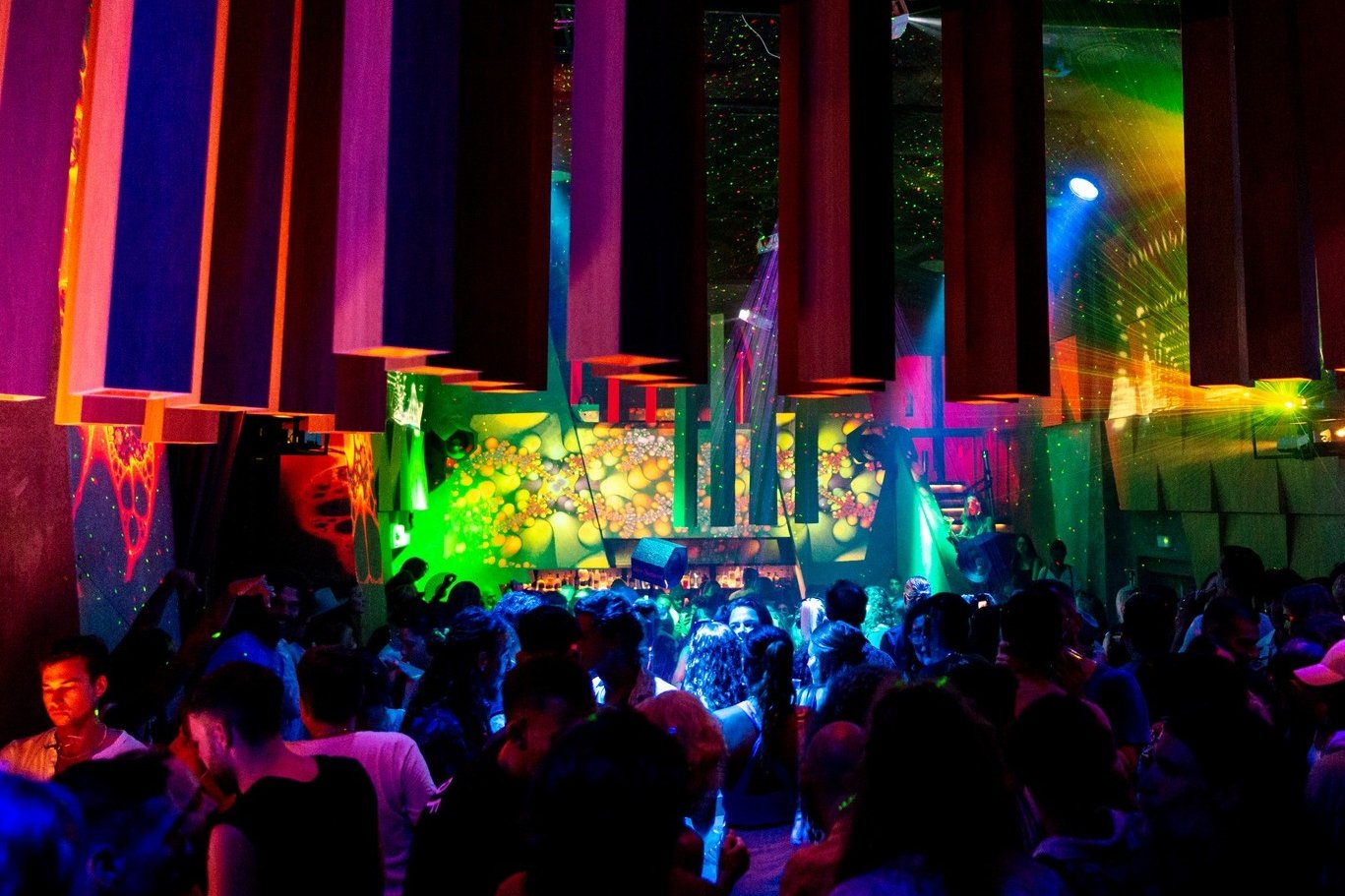 Dance floor with colorful panels hanging from the ceiling, a wall of green and yellow projections, lasers, and the crowd dancing