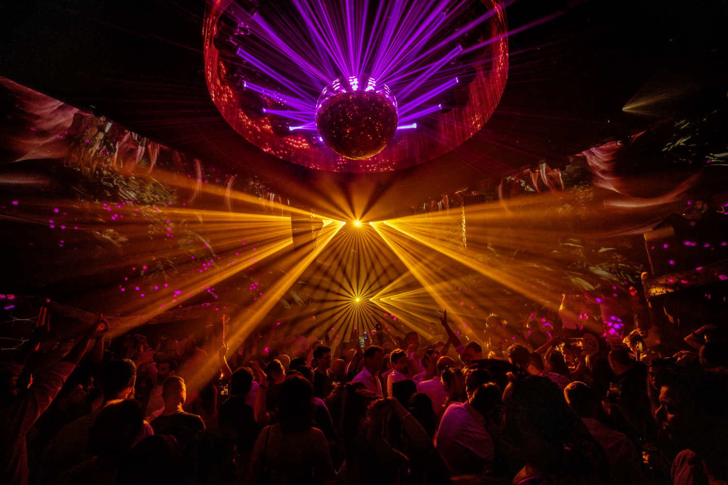 Packed Chinois pista, with a mirror ball beneath a ring of red lights and yellow laser beams sweeping over the crowd with arms raised