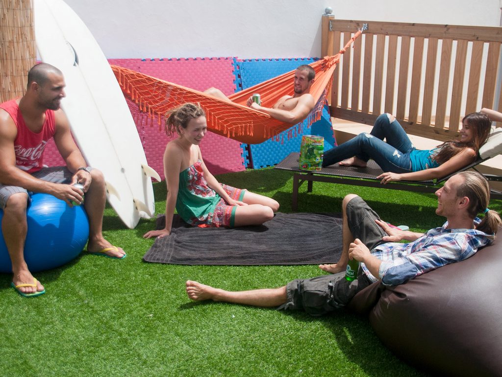 Travellers relaxing on an artificial-grass terrace, one in an orange hammock, with a surfboard against the wall