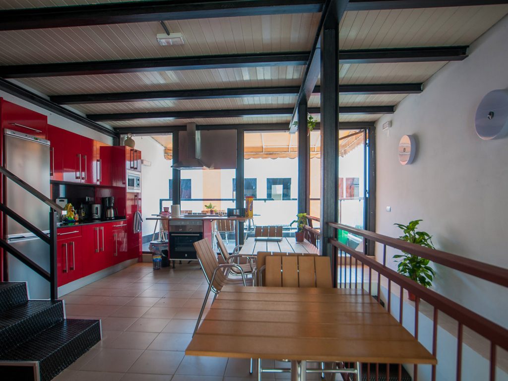 Open-plan kitchen with red cabinets and wooden dining tables beside a glass wall onto the terrace