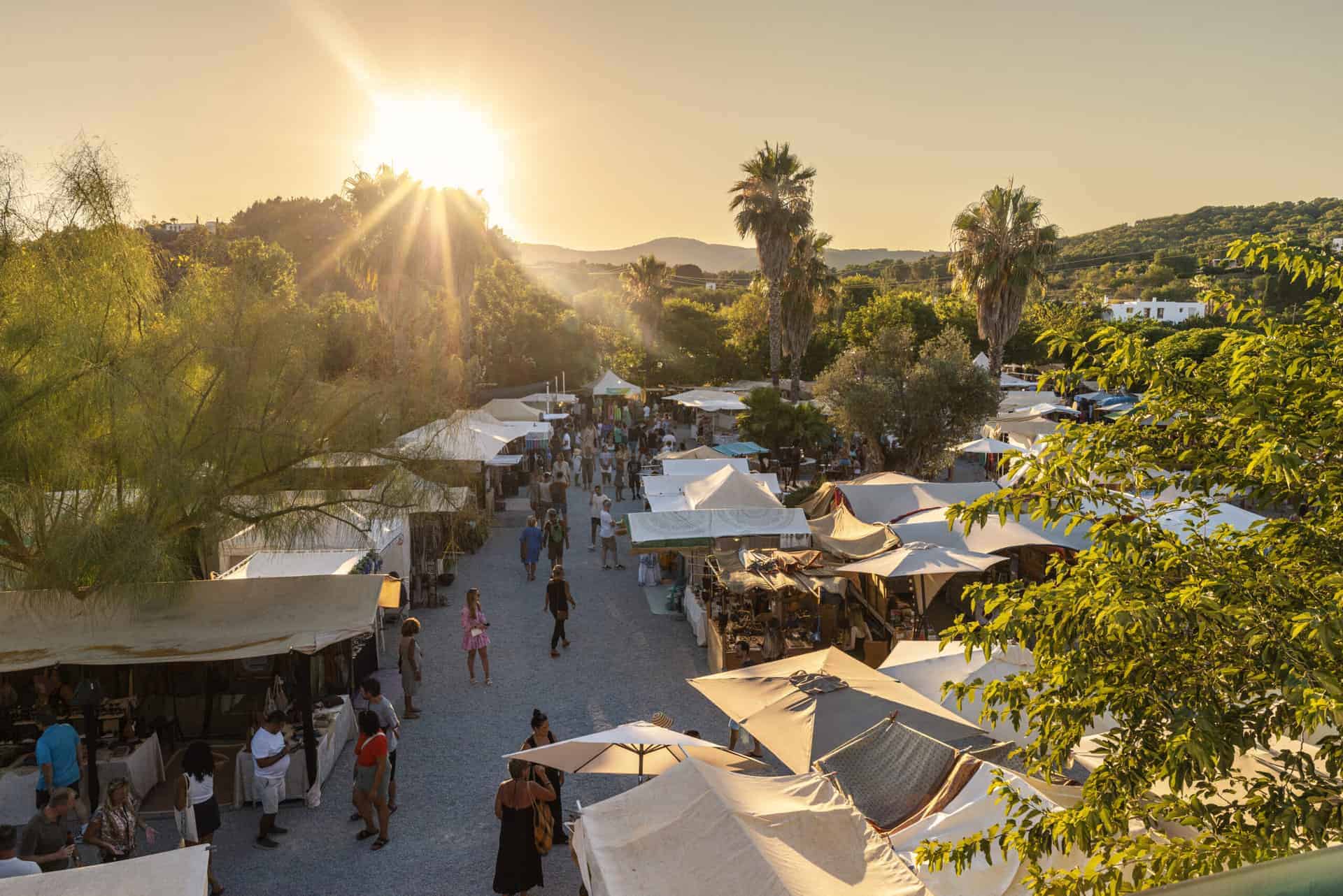 Street of white-canopied market stalls seen from above, with people strolling, palm trees, and the low sun setting behind the trees