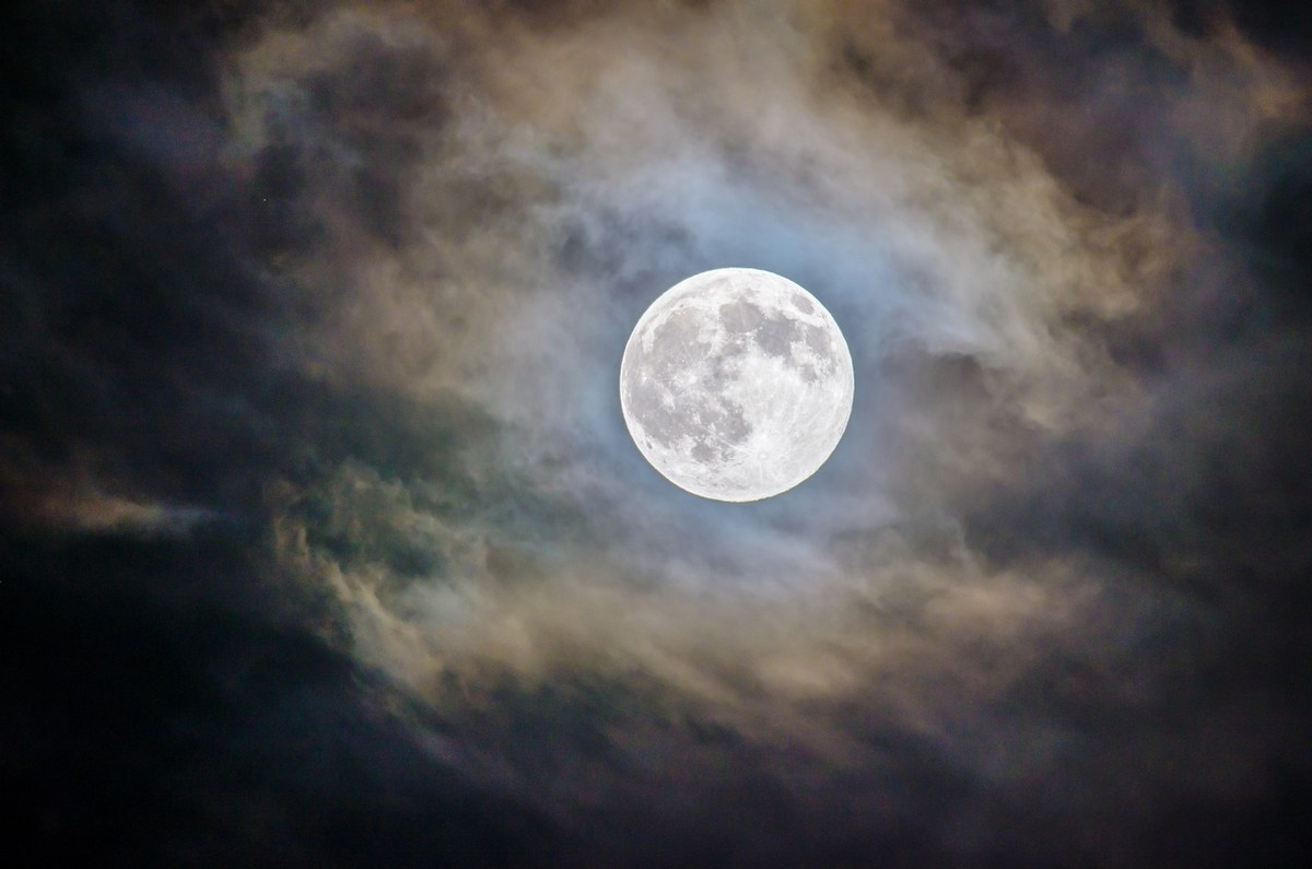 Full moon at night, surrounded by clouds lit up in shades of blue and gold